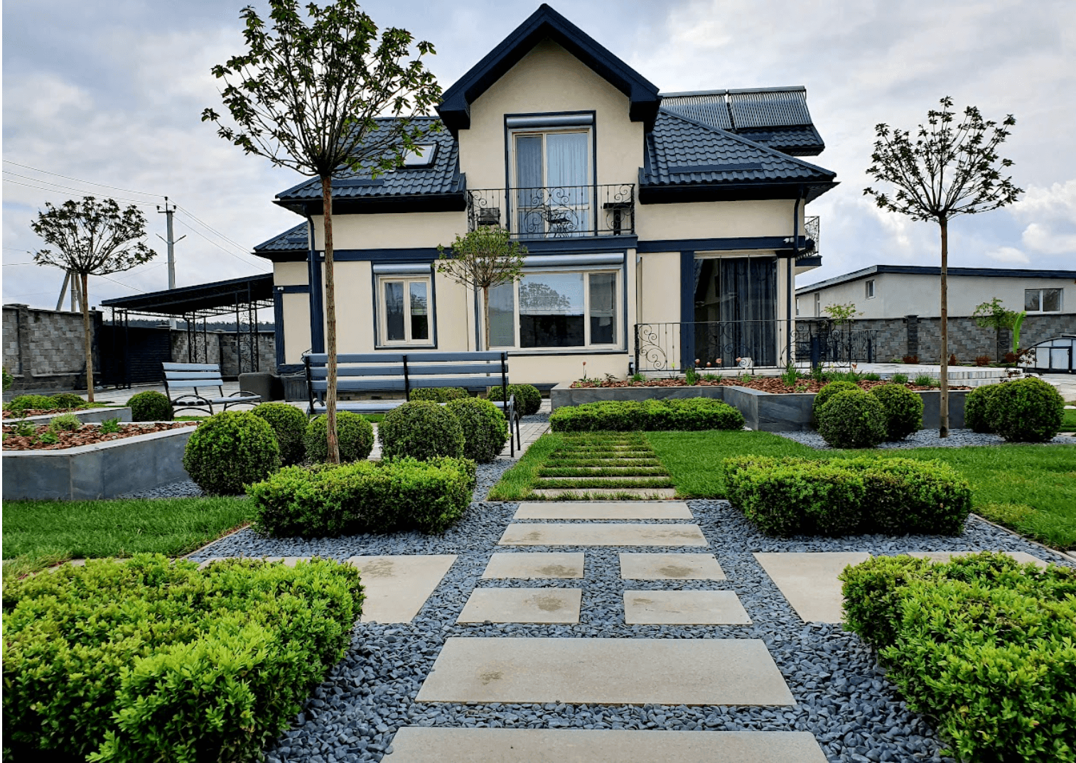 Modern beige house with dark trim and a manicured garden featuring spherical bushes and paths.