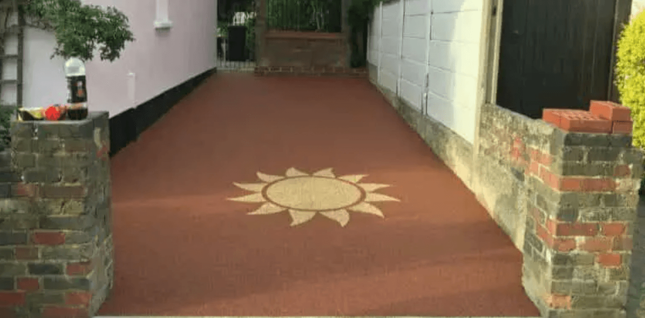 Reddish-brown driveway with a decorative sun emblem, bordered by a house and brick walls.